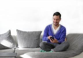 lächelnder Mann, der mit dem Smartphone umgeht, während er auf der Couch sitzt foto