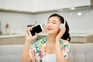 schöne, emotional erfreute junge Frau auf dem Sofa zu Hause, die Musik mit Kopfhörern hört, die das Handy singen halten. foto