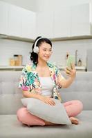 Porträt schöne junge asiatische Frau verwendet ein intelligentes Mobiltelefon mit Kopfhörer, um Musik auf dem Sofa im Wohnzimmerbereich zu hören foto