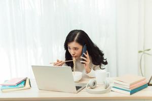 selbstbewusste lächelnde attraktive junge asiatische unternehmerin, die mittagspause macht, während sie am schreibtisch sitzt und ein handy benutzt foto