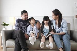 asiatische thailändische familie, erwachsener vater, mutter und kinder glück zu hause leben entspannende aktivitäten und gemeinsames lesen von büchern, freizeit auf dem sofa im weißen zimmerhaus, schönes wochenende, häuslicher lebensstil des wohlbefindens. foto