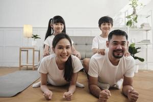 glückliches wohlbefinden asiatische thailändische familie, kinder spielen und necken ihre eltern beim yoga-fitnesstraining und gesundheitsübungen zusammen im weißen wohnzimmer, häuslicher lebensstil, wochenendaktivität. foto