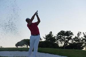 Golfspieler, der einen Sandbunker schlägt, der bei Sonnenuntergang erschossen wird foto