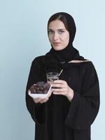 Moderne muslimische Frau in Abaya, die eine Dattelfrucht und ein Glas Wasser vor sich hält foto