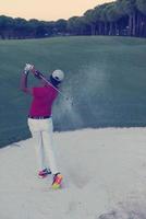 Golfspieler, der einen Sandbunker schlägt, der bei Sonnenuntergang erschossen wird foto