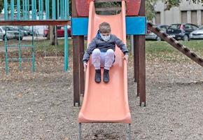 kleiner junge mit schützender gesichtsmaske, die auf dem spielplatz rutscht. foto