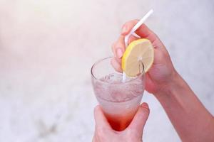 Frauenhände, die ein Glas eisgekühltes Sommergetränk mit in Scheiben geschnittener Zitrone am Sandstrand halten foto