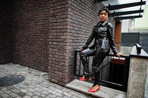 Modische, schöne Afroamerikanerin, die in schwarzer Lederjacke und Hose auf der Straße posiert. foto