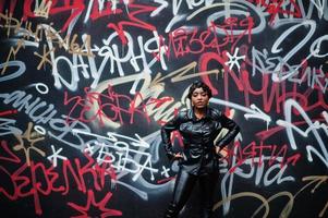 modische schöne afroamerikanische frau, die in schwarzer lederjacke und hose auf der straße gegen graffitiwand posiert.