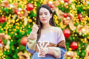 asiatische schöne dame in einem bunten pullover steht glücklich. in seiner hand hielt er ein licht vor dem weihnachtsbaum. mit bokeh als hintergrund im thema weihnachten und neujahrsfeiern foto