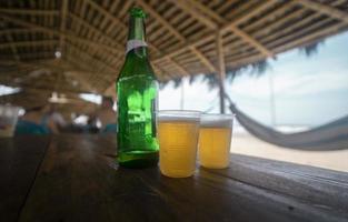 Zwei Plastikgläser mit Bier und eine grüne Flasche auf einem Holztisch in einem Restaurant am Strand mit Bambusdach und Hängematten an den Seiten foto