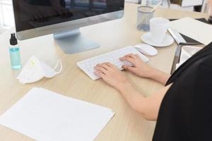 eine geschäftsfrau, die in einem büro arbeitet und auf einer tastaturtasche tippt, während eine alkoholsprühflasche und eine n95-maske auf ihrem schreibtisch stehen.