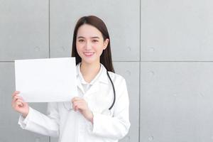 asiatische ärztin, die einen medizinischen mantel trägt, hält und zeigt weißes papier, um etwas im gesundheitskonzept zu präsentieren. foto