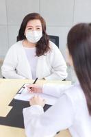 asiatische ärztin zeichnet symptome des patienten während der diagnose auf dem dokument auf, beide tragen im krankenhaus eine gesichtsmaske. foto