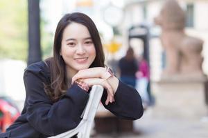 porträt asiatische schöne frau in dunkelblauem langarmhemd sitzt und lächelt auf stuhl in einem städtischen park im freien. foto