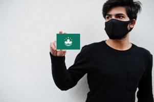 asiatischer mann trägt ganz schwarz mit gesichtsmaske und hält macau-flagge in der hand isoliert auf weißem hintergrund. Coronavirus-Länderkonzept. foto