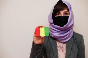 porträt einer jungen muslimischen frau, die formelle kleidung trägt, gesichtsmaske und hijab-kopftuch schützt, guinea-flaggenkarte vor isoliertem hintergrund hält. Coronavirus-Länderkonzept. foto