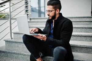 nahöstlicher unternehmer trägt schwarzen mantel und blaues hemd, brille gegen bürogebäude mit laptop. foto