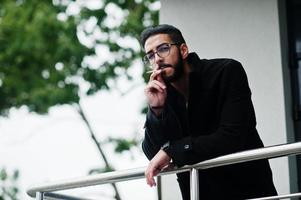 nahöstlicher unternehmer trägt schwarzen mantel und blaues hemd, brille gegen rauchende zigarette des bürogebäudes. foto