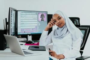 Junge afroamerikanische moderne muslimische Geschäftsfrau mit Schal an einem kreativen, hellen Büroarbeitsplatz mit großem Bildschirm. foto