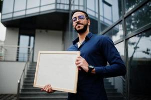 nahöstlicher mann trägt blaues hemd, brille, hält weißes leeres brett. foto