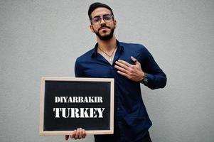 arabischer mann trägt blaues hemd und brille hält brett mit diyarbakir-türkei-inschrift. größte städte im islamischen weltkonzept. foto