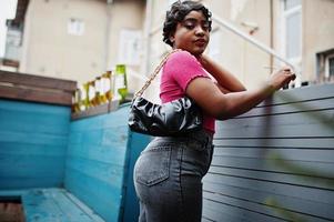 urbane junge afrikanisch-amerikanische frau in rosa top und grauen jeans und sonnenbrille mit handtasche. afro mode schicke frauen. foto