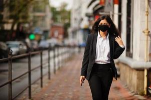 wunderschöne indische frau trägt eine formelle und schwarze gesichtsmaske und posiert während der covid pandemia auf der straße, mit dem handy zur hand. foto