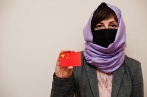 porträt einer jungen muslimischen frau, die formelle kleidung trägt, gesichtsmaske und hijab-kopftuch schützt, china-flaggenkarte vor isoliertem hintergrund hält. Coronavirus-Länderkonzept. foto