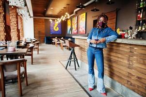 stilvolle afroamerikanische frau mit dreadlocks afro-haar, trägt jeansjacke und gesichtsschutzmaske im restaurant, hält handy. neues normales leben nach der coronavirus-epidemie. foto