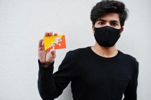 asiatischer mann trägt ganz schwarz mit gesichtsmaske und hält bhutan-flagge in der hand isoliert auf weißem hintergrund. Coronavirus-Länderkonzept. foto