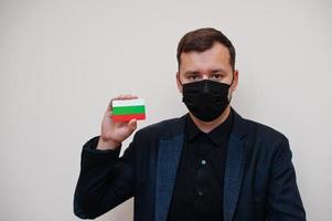 europäischer mann trägt schwarze formale und schützt gesichtsmaske, hält bulgarien-flaggenkarte lokalisiert auf weißem hintergrund. europa coronavirus covid country konzept. foto