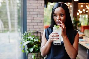 modische feministische afroamerikanerin in schwarzem t-shirt und shorts, posiert im restaurant mit limonadenglas. foto