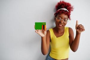 afrikanische frau mit afrohaar, trägt gelbes unterhemd und brille, hält sambia-flagge isoliert auf weißem hintergrund, zeigt daumen hoch. foto