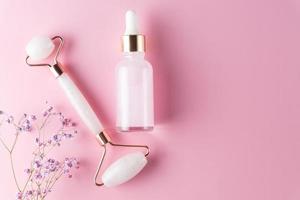 Massage-Quarzrolle für das Gesicht aus Naturstein mit Serumöl in transparenter Glasflasche mit Gypsophila-Blüten auf rosafarbenem Hintergrund.