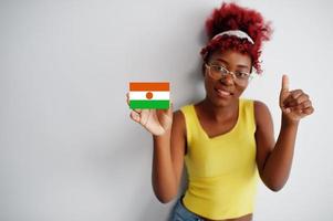 afrikanische frau mit afro-haar, trägt gelbes unterhemd und brille, hält niger-flagge isoliert auf weißem hintergrund, zeigt daumen hoch. foto