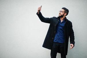 Unternehmer aus dem Nahen Osten tragen einen schwarzen Mantel und ein blaues Hemd, eine Brille steht an einer weißen Wand, raucht eine Zigarette und macht ein Selfie per Telefon. foto