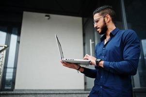 nahöstlicher unternehmer trägt blaues hemd, brille mit laptop gegen bürogebäude. foto