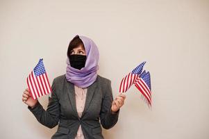 porträt einer jungen muslimischen frau, die formelle kleidung trägt, gesichtsmaske und hijab-kopftuch schützt, usa-flaggen in den händen vor isoliertem hintergrund hält. Coronavirus-Länderkonzept. foto