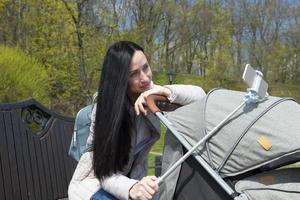 junge schöne mutter in dunklen haaren, eine frau mit einem süßen baby in einem kinderwagen macht ein selfie vom telefon. foto