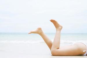 bräunen sonnengebräunte junge frau im bikini mit strohhüten, die an einem tropischen strand auf weißem sand liegen, der sich ein schlankes bein ausdehnt, blaues meer im hintergrund. sommerferien modeurlaub reisekonzept. foto