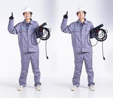 in voller länge 20er jahre junge mix renningenieur elektrikerin, zeige finger in die luft, trage graue uniform. unternehmer weiblich halten elektrische stecker kabel lächeln glücklich über weißem hintergrund isoliert foto