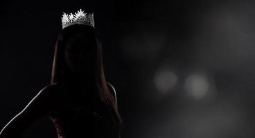 Porträt des Miss-Schönheitswettbewerbs im Pailletten-Abend-Ballkleid, langes Kleid mit funkelnder, heller Diamantkrone, Silhouette, zurückhaltende Belichtung mit Vorhang, Studiobeleuchtung, dunkler Hintergrund, dramatisch foto
