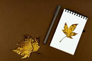 Herbstgeschäftskonzept - leeres, ringgebundenes Notizbuch mit goldenem Ahornblatt und Stift auf braunem Hintergrund mit Kopierraum foto