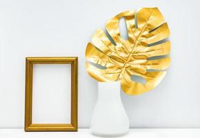 gold-weißes innenmodelldesign. leerer fotorahmen und monsterblatt im weißen vase auf weißem wandhintergrund. trendiges Design foto