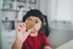 asiatisches baby, das ein rotes t-shirt trägt, das goldene bitcoin-kryptowährung blockchain auf holztischschreibtisch im wohnzimmer zu hause hält. Krypto-Währungs-Blockchain-Konzept zur Einsparung von Anlagevermögen. foto