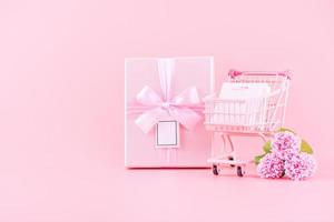 muttertag urlaubsgeschenk designkonzept, rosa nelkenblumenstrauß mit umwickelter schachtel, einkaufswagen, tasche, einzeln auf hellrosa hintergrund, kopierraum. foto
