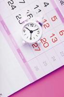 Weißer Wecker und Kalender auf einem rosa Desktop-Hintergrund. foto