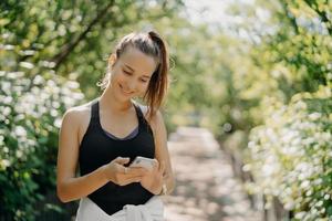 Außenaufnahme einer glücklichen, athletischen jungen Frau verwendet eine Smartphone-Sport-Tracker-Anwendung, die ein modernes Gerät für Gesundheit und Fitness verwendet, das sich mit Trainingsposen vor verschwommenem Naturhintergrund auskennt foto