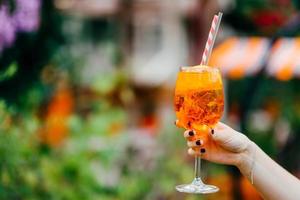 schuss der frauenhand mit maniküre halten frisches sommergetränk im weinglas. orangefarbener kalter Cocktail mit Eis, den Sie vor verschwommenem Hintergrund trinken können foto